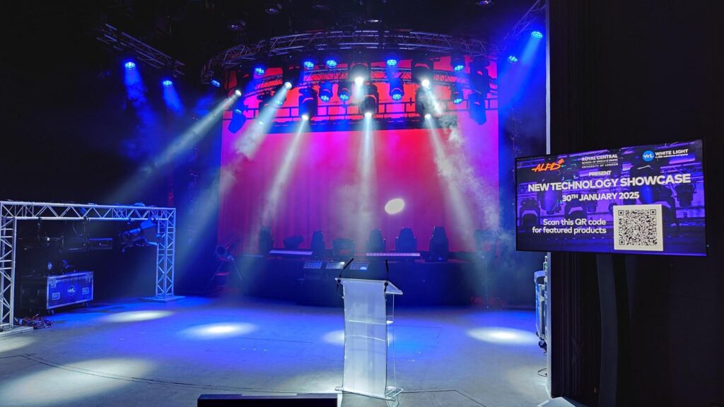 A stage lit with moving head lights, piercing through haze. An empty podium, truss and display showing the events date can be seen.